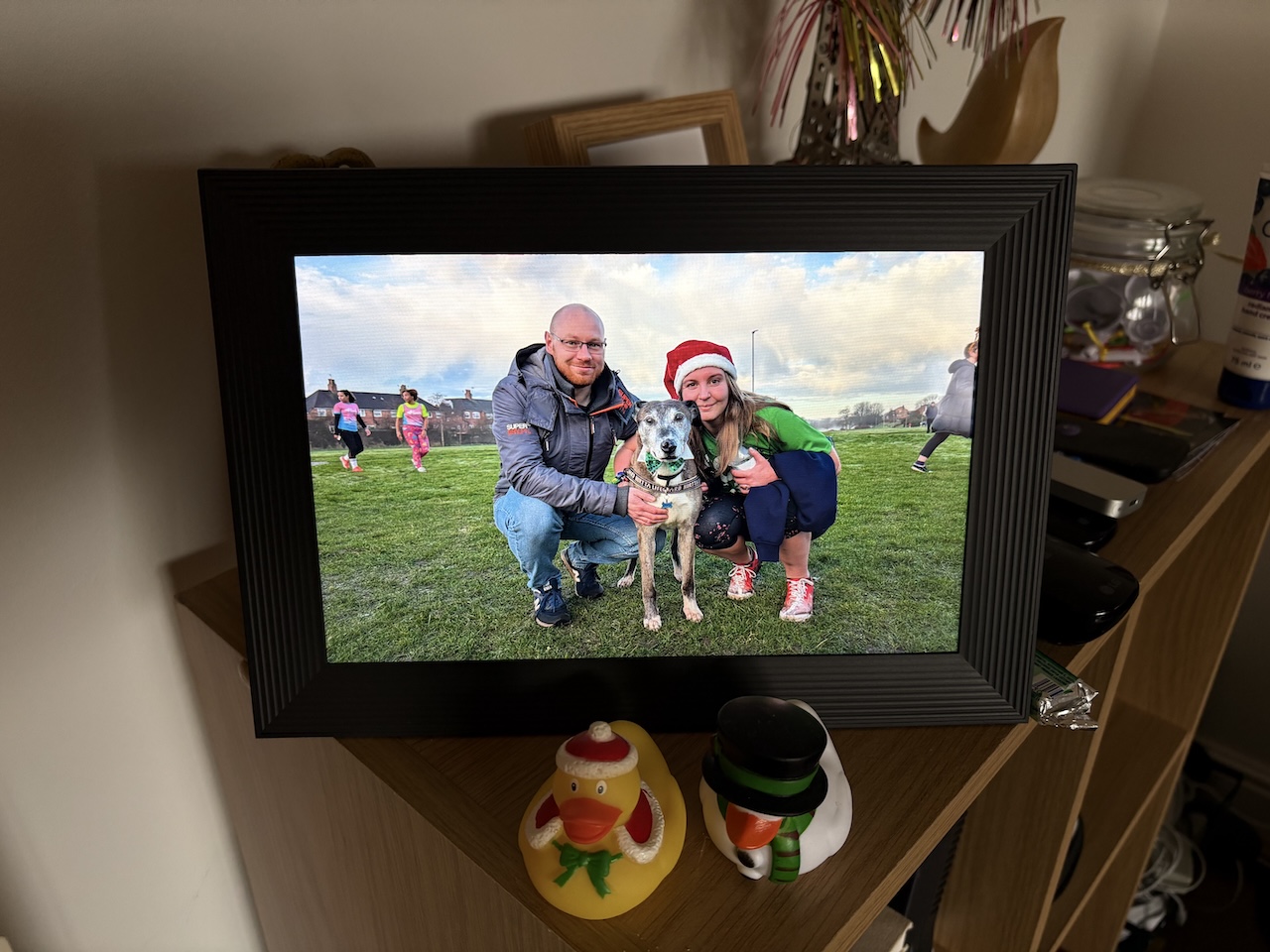 A photo of the Aura Carver digital photo frame, showing a photo of me, my wife, and our dog at a Christmas Day Parkrun. The frame is perched atop an admittedly overcrowded half-height bookcase. There are two Christmas-themed rubber ducks in front of it, though I'm not sure why.